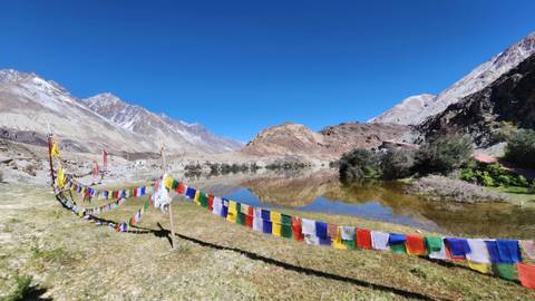 Colourful prayer flags line a lakeside meadow with reflections of barren peaks and clear blue sky in still water.