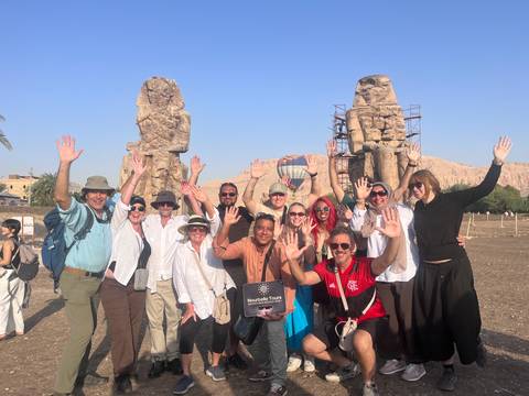 Tour group cheers with raised hands in front of the Colossi of Memnon with a balloon in the background.