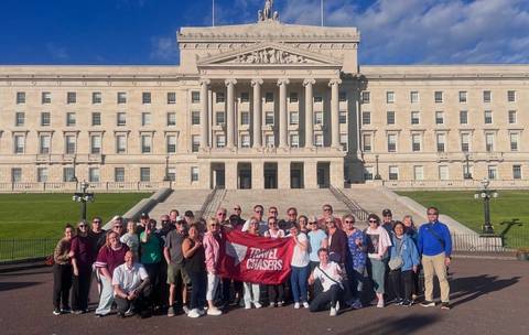 Large tour group posing with a red banner on the steps of a neoclassical government building under blue skies