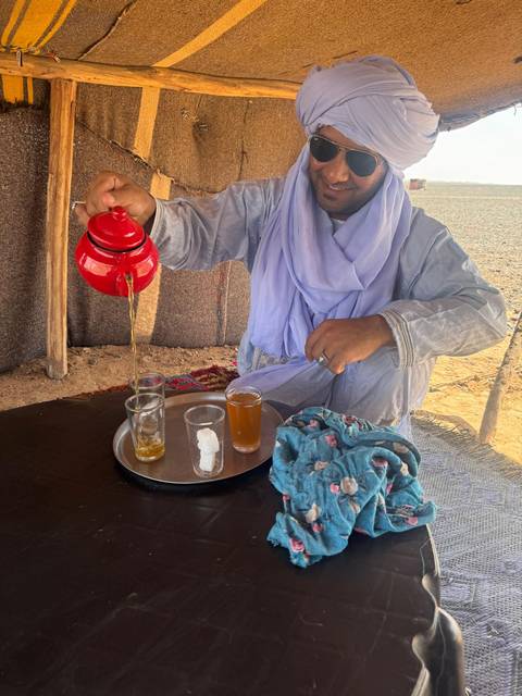 Berber host pouring steaming mint tea from red teapot into glasses inside a desert tent