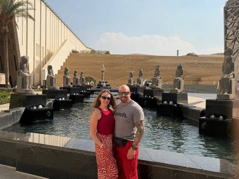 Couple posing by modern fountains lined with pharaonic statues outside a grand museum entrance.