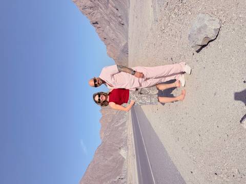 Travelers stand barefoot on a lonely desert highway flanked by rugged brown mountains under clear blue skies.