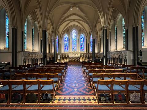 Elegant Gothic cathedral nave with patterned tile floor, wooden chairs, and colorful stained glass windows.