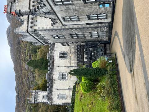 Gray stone Victorian-era castle nestled at the base of a wooded mountain slope.