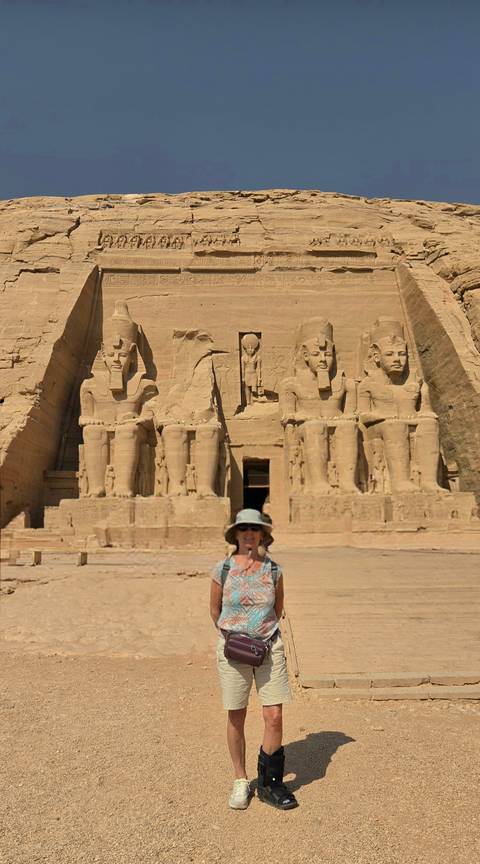 Front façade of Abu Simbel with four colossal seated statues; a tourist wearing a sun hat poses in the foreground.