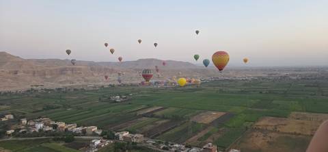 Dozens of colorful hot-air balloons float above fertile green fields and desert cliffs near Luxor at dawn.