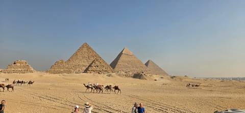 Wide desert panorama of the three main pyramids with camel caravans and tourists traversing the sandy foreground.