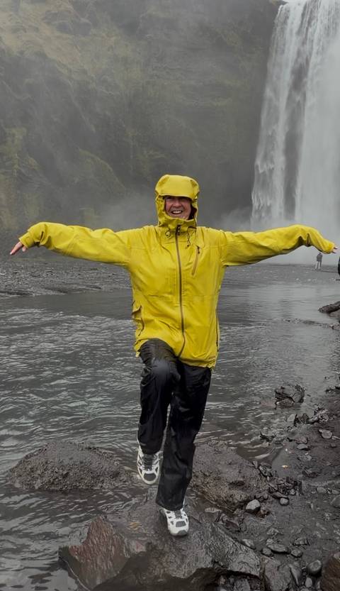 Smiling traveller in a bright yellow rain jacket throws out her arms beside a misty waterfall.