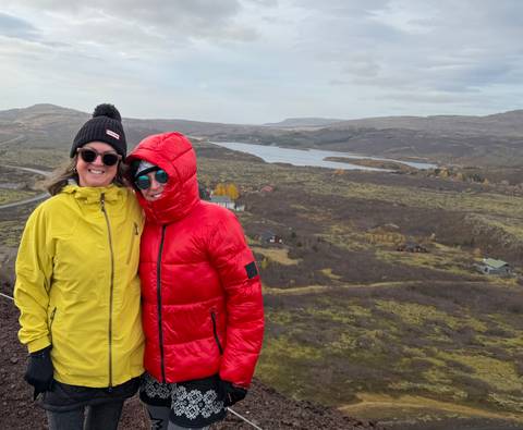 Two friends in bright jackets smile with a sweeping lake and tundra landscape behind them.