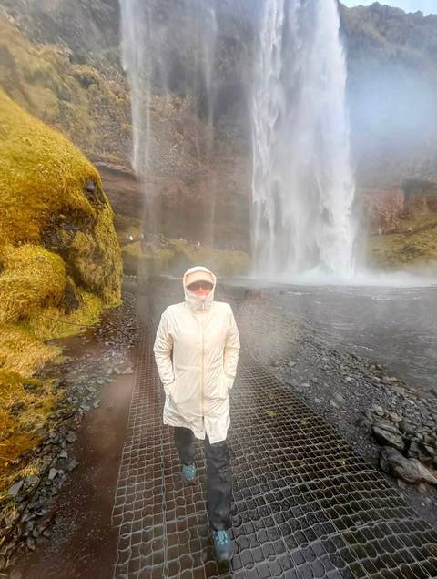 Rain-coated traveller stands on a wet metal grate beneath a powerful waterfall’s spray.