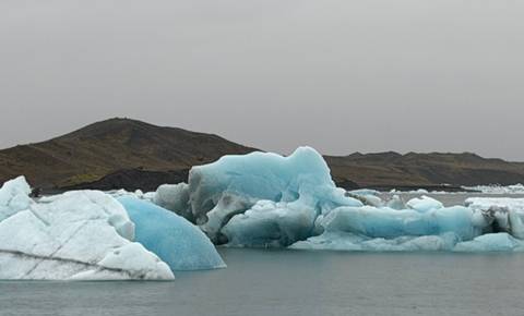 Pale-blue icebergs float in a still glacial lagoon beneath a grey sky.