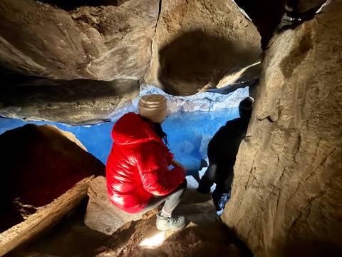 Travellers crouch at the mouth of a blue-lit lava cave surrounded by rough stone walls.