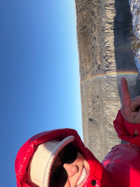 A pointing hand foregrounds a narrow rainbow rising from a deep canyon under blue skies.