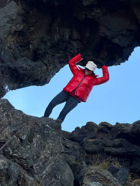 Traveller in red jacket climbs through a natural lava arch against a clear blue sky.