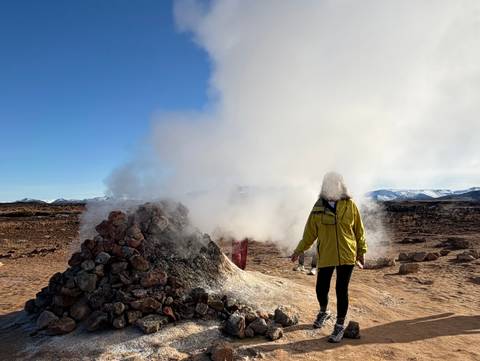Steam billows from a rocky fumarole on a barren geothermal plain as visitors explore.