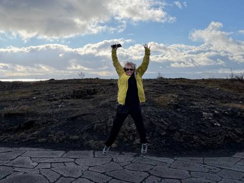 Person in yellow jacket jumps with arms raised on a barren lava plain under dramatic clouds.