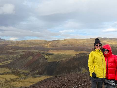 Two friends in colourful jackets stand on the rim of a volcanic crater under a cloudy sky.