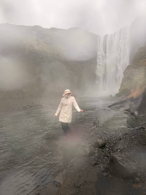 Traveller in raincoat stands in heavy mist before a tall waterfall, surroundings blurred with spray.
