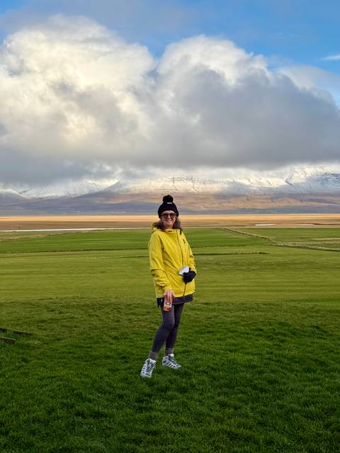 Smiling traveller in yellow jacket stands on rolling green fields with snow-capped peaks behind.
