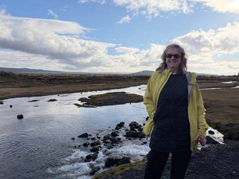 Traveller in yellow jacket stands beside a reflective river under a patchy cloud sky.