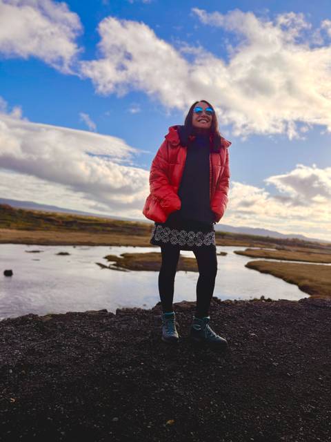 Partially framed figure in red jacket stands on rocky bank with blurred countryside backdrop.