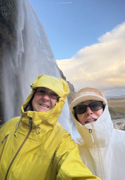 Close selfie of two travellers in rain gear beside a tall waterfall curtain.