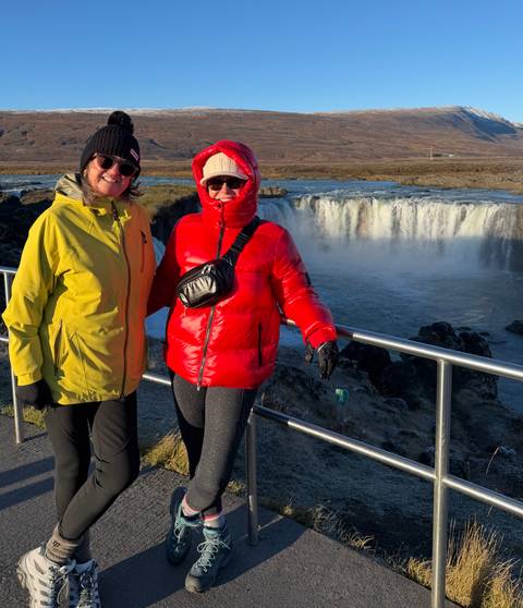 Two warmly dressed women pose by a railing overlooking Godafoss waterfall in bright sunlight.