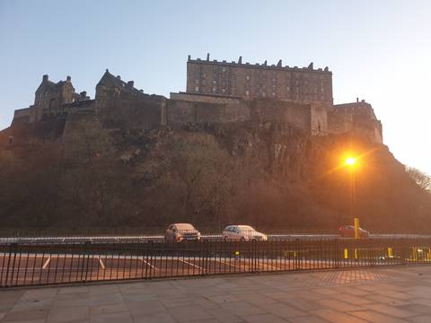 Edinburgh Castle crowned on a rugged volcanic hill at dusk with a streetlamp glowing in the foreground and parked cars below the ramparts.