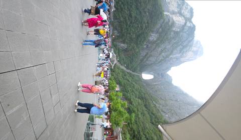 Wide plaza full of tourists looking toward the giant natural arch of Tianmen Mountain rising steeply behind a long staircase.