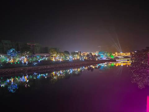 Colorful riverside light display reflecting in calm water against a night sky with searchlights beaming upward.