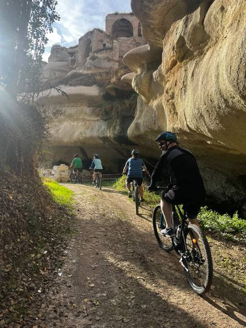 Cyclists ride single file toward a cave entrance carved in soft Cappadocian rock, sunlight streaming through trees.