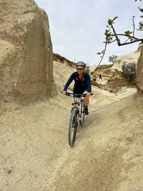 Smiling woman navigates a narrow sandy valley on her mountain bike amid pale tufa formations.