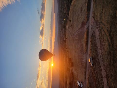 Silhouette of a hot-air balloon floating above Cappadocia at sunrise with golden sun on the horizon and a lone vehicle on a dirt track below.
