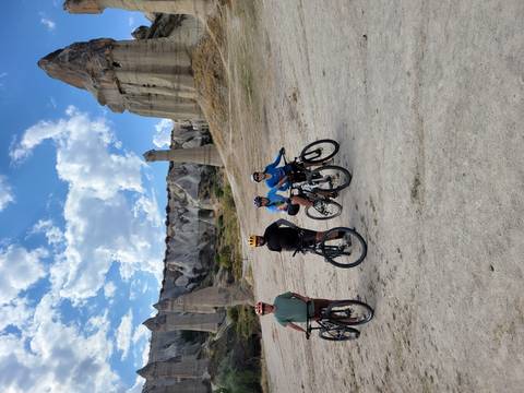 Cyclists with bikes pose in front of Cappadocia’s famous phallic rock formations in Love Valley under scattered clouds.