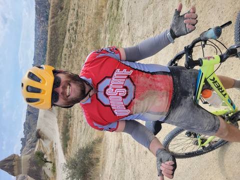 Dust-covered cyclist in Ohio State jersey smiles holding his mountain bike after a ride on Cappadocian trails.