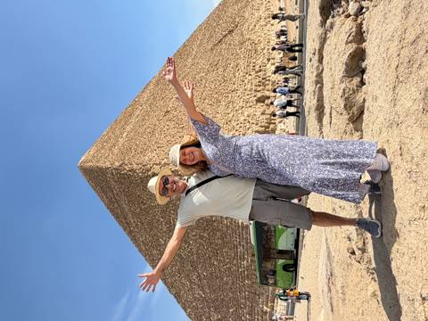 Smiling couple with arms outstretched stand before the Great Pyramid of Giza on a clear blue day.