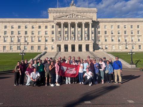 A large tour group poses with a Travel Chasers banner on the steps of Stormont Parliament in Belfast.