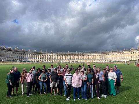 A tour group smiles before the sweeping Georgian arc of Bath’s Royal Crescent under dramatic skies.