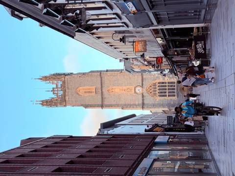 A tall Gothic church tower rises above a pedestrian street lined with shops and cyclists.