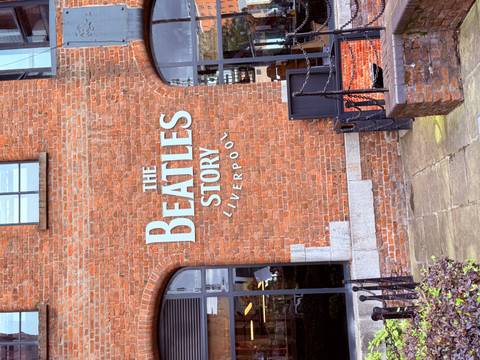 Red-brick warehouse wall featuring a large sign reading 'The Beatles Story Liverpool' with visitors walking past.