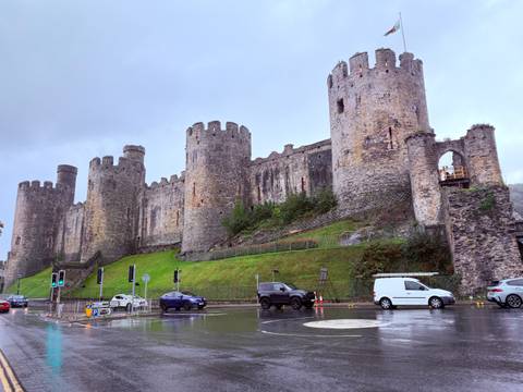 Massive medieval stone castle with circular towers rising above a rainy street and lush green embankment.