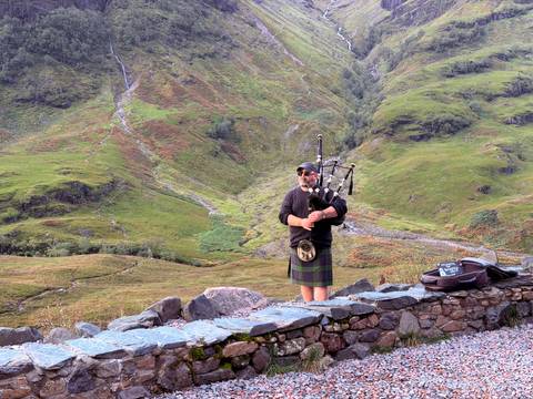 Traditional Scottish bagpiper in kilt performing on a stone wall with verdant Highlands backdrop.