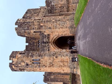 Visitors enter a medieval stone gateway of a large castle surrounded by green lawns and a paved path.