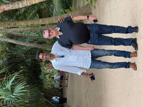 Two men smile for the camera while strolling along a shady path lined with tropical palms.