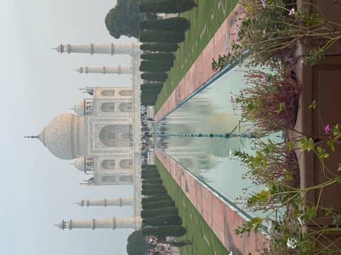 The Taj Mahal reflected in its central pool with rows of cypress trees and visitors in the distance.