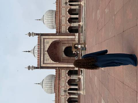 A woman in a long abaya stands before the grand red-sandstone arches of Jama Masjid in Delhi.