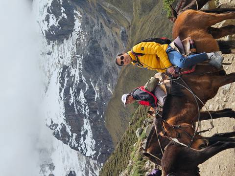 Two trekkers on horseback pause before snow-covered Andean mountains shrouded in mist.