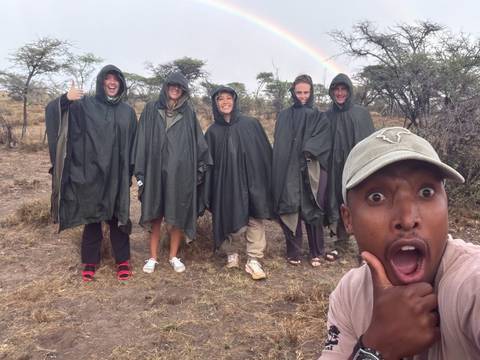 Group of travellers wearing ponchos posing cheerfully on a safari plain with a rainbow overhead.