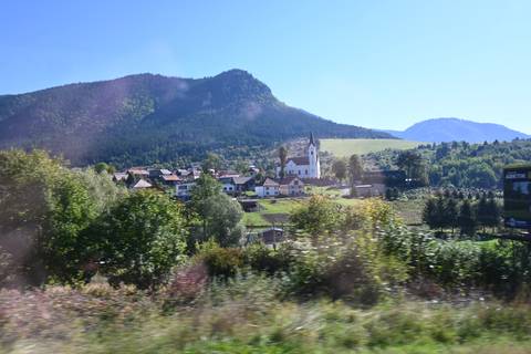 Blurred view from a moving vehicle of a small alpine village beneath forested mountains.