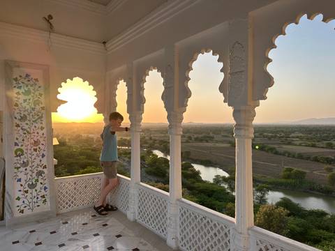 Boy leans on a white lattice balcony enjoying a golden sunset over a winding river and green countryside.
